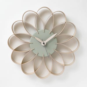 Peut inclure: Une horloge murale de couleur vert clair et bois naturel avec un design en forme de fleur. L'horloge a un cadran rond avec des aiguilles noires et un fond vert clair.