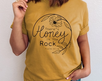 Może przedstawiać: Żółta koszulka w kolorze musztardowym z czarnym wzorem kwiatowym i tekstem "There's Honey in the Rock Psalms 81:16".