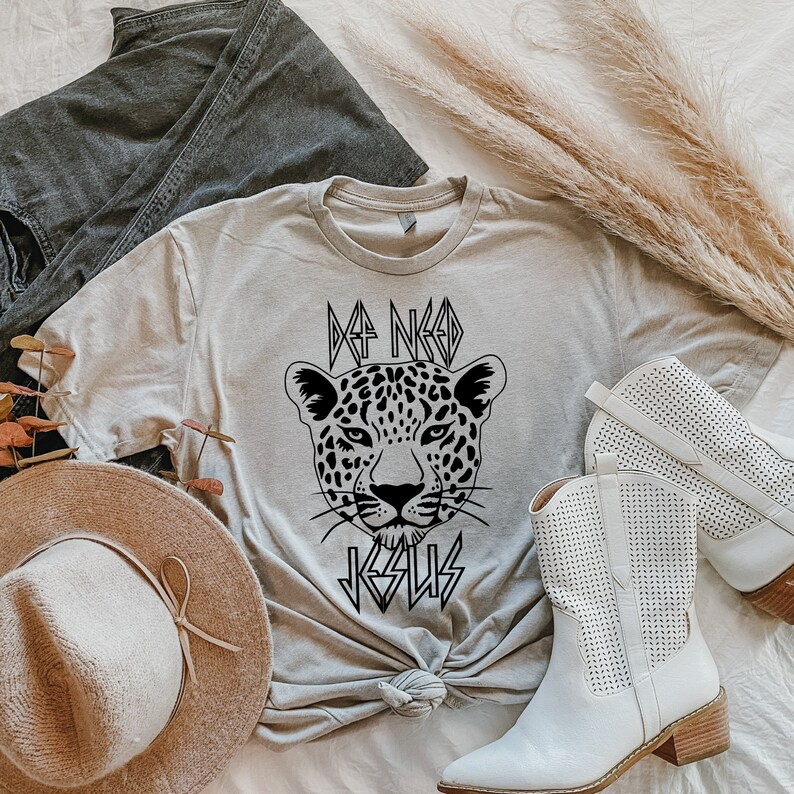 May include: A light gray t-shirt with a black graphic of a leopard's face and the text "I NEED JESUS" in a bold, stylized font.