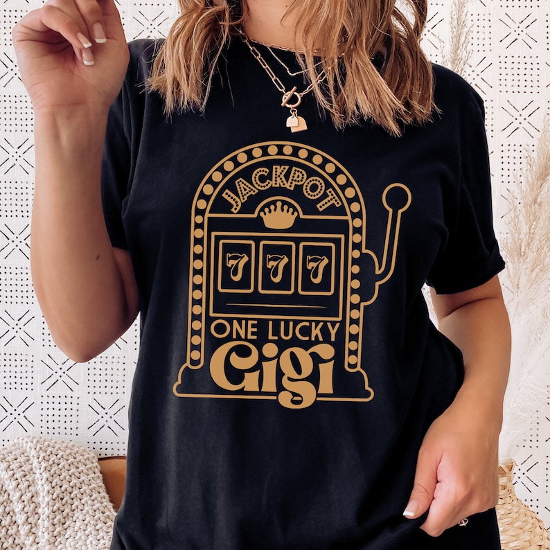 Op de afbeelding: Zwart T-shirt met een gouden afbeelding van een gokautomaat met de tekst "JACKPOT" en "ONE LUCKY Gigi".