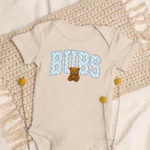 Puede incluir: Un body de bebé beige claro con la palabra "BUBS" en letras a cuadros azules y un gráfico de oso de peluche marrón. El body está hecho de material suave y está diseñado para bebés.