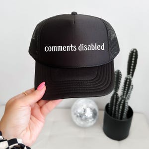 May include: Black trucker hat with a mesh back and the words "comments disabled" in white. The hat is held up by a person's hand. A small disco ball and a potted cactus are in the background.