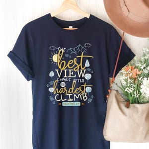 以下が含まれることがあります： 紺色のTシャツ。金と白で「The best view comes after the hardest climb」と書かれています。デザインには太陽、山、木、足跡が含まれています。「ガラテヤ人への手紙6:9」のテキストも含まれています。