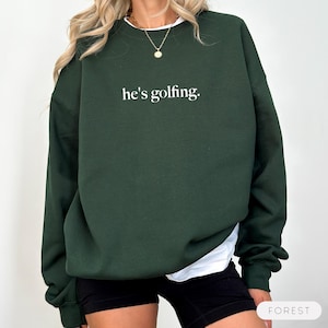 May include: A dark green sweatshirt with the text "he's golfing." in white. The sweatshirt is a solid color and has long sleeves. The model is wearing a white shirt underneath and black shorts. The word "FOREST" is in the bottom right corner.