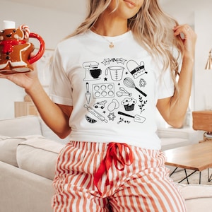 Pode incluir: Camiseta branca com uma ilustração em preto e branco de várias ferramentas e ingredientes para cozedura. A ilustração inclui uma batedeira, uma chávena de medição, um rolo, cortadores de biscoitos e um cupcake.