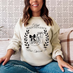May include: Cream-colored sweatshirt with the text "High Maintenance But Worth It" in a black script font. The design includes a wreath, a wrapped gift, and a jar. The sweatshirt is a casual style.