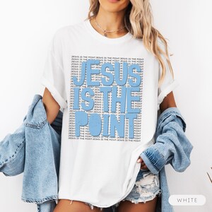 May include: White t-shirt featuring the text "JESUS IS THE POINT" in light blue, with the phrase "JESUS IS THE POINT" repeated in a smaller font. The shirt has a relaxed fit.