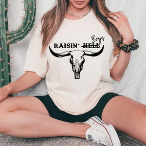 Op de afbeelding: Een wit T-shirt met een zwarte stieren schedel afbeelding en de tekst "Boys Raisin' Hell".