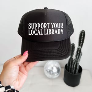 Peut inclure: Casquette de camionneur noire avec le texte blanc "SUPPORT YOUR LOCAL LIBRARY". Le chapeau a un dos en filet et une visière incurvée. Le chapeau est tenu par une personne.