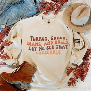 以下が含まれることがあります： レトロなフォントで「TURKEY, GRAVY, BEANS, AND ROLLS, LET ME SEE THAT CASSEROLE」と書かれたベージュのスウェットシャツです。