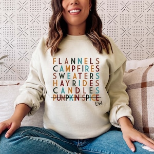 Puede incluir: Una sudadera de color crema con un estampado gráfico de las palabras "Flannels, Campfires, Sweaters, Hayrides, Candles, Pumpkin Spice" con una línea que atraviesa "Pumpkin Spice" y la palabra "Ew." impresa debajo.