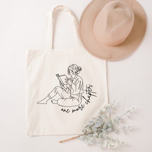 Op de afbeelding: Een witte canvas tote bag met een zwarte lijntekening van een persoon die een boek leest. De tekst "one more chapter" is geschreven in een cursief schrift onder de tekening.