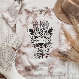 May include: A white and brown tie-dye sweatshirt with a black and white leopard print graphic and the text "I Need Jesus" in a bold, stylized font.