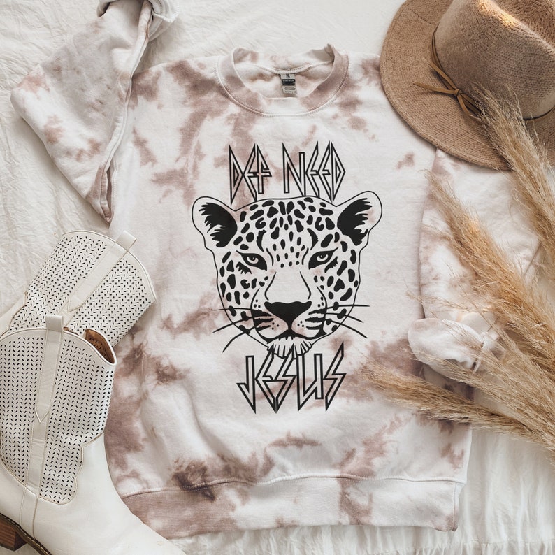May include: A white and brown tie-dye sweatshirt with a black and white leopard print graphic and the text "I Need Jesus" in a bold, stylized font.