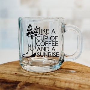 May include: A clear glass mug with a black silhouette of a cowboy boot with flowers in the top and the text "Like a cup of coffee and a sunrise".
