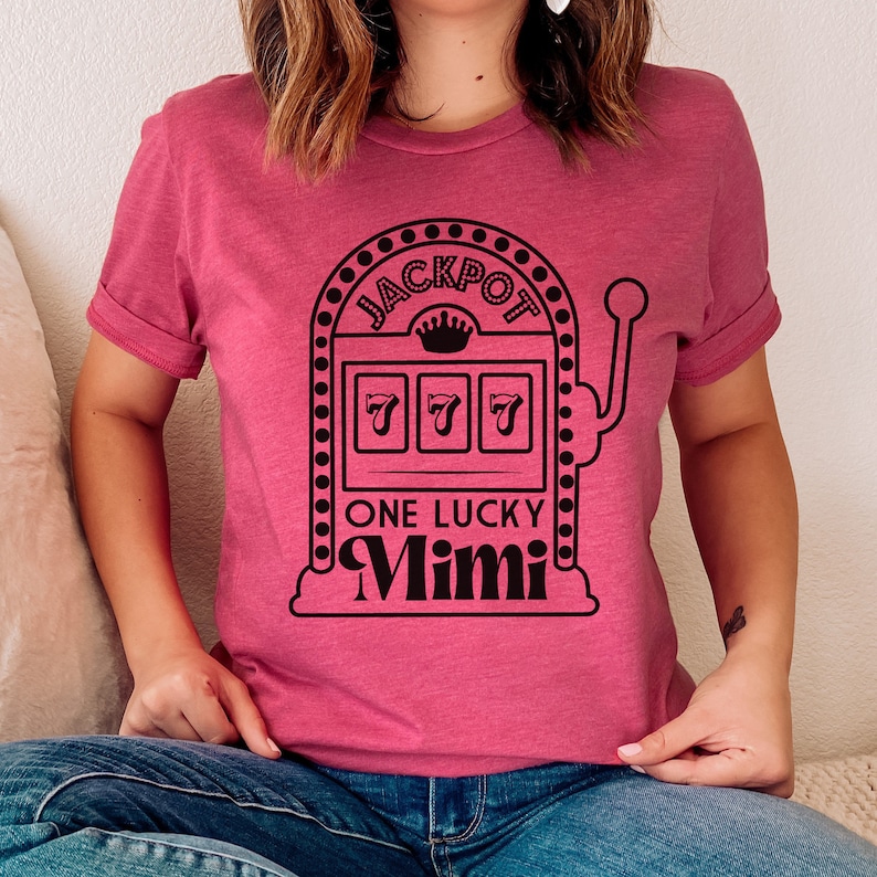 Op de afbeelding: Een roze T-shirt met een zwarte afbeelding van een gokautomaat met de tekst "JACKPOT" boven de rollen en "ONE LUCKY Mimi" onder de rollen. De rollen tonen drie zevens.