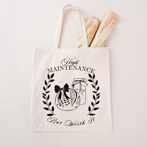 May include: A white canvas tote bag with black text that reads "High Maintenance But Worth It". The bag features a black and white illustration of a jar with a bow and a wreath of leaves.