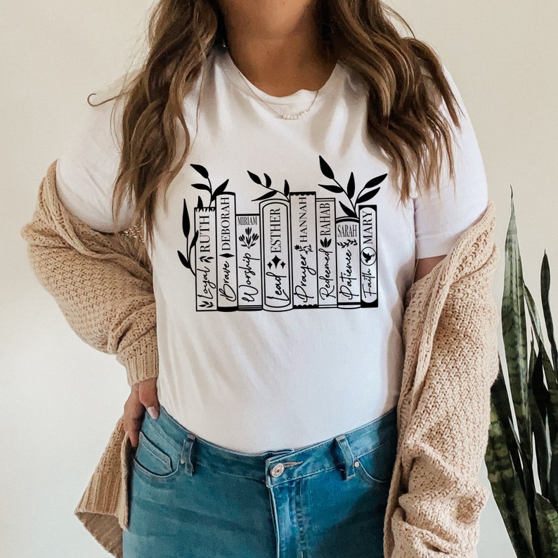 May include: White t-shirt with a black graphic of a bookshelf with books that have names of women from the Bible. The books are "Loyal Ruth", "Brave Deborah", "Worship Miriam", "Lead Esther", "Prayer Hannah", "Refined Rahab", "Patience Sarah", and "Faith Mary".