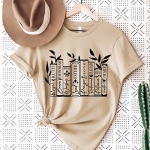 May include: A beige t-shirt with a black graphic of a bookshelf with books that have names of women from the Bible. The books are labeled: Loyal Ruth, Brave Deborah, Worship Miriam, Lead Esther, Prayer Hannah, Redeemed Rahab, Patience Sarah, Faith Mary.