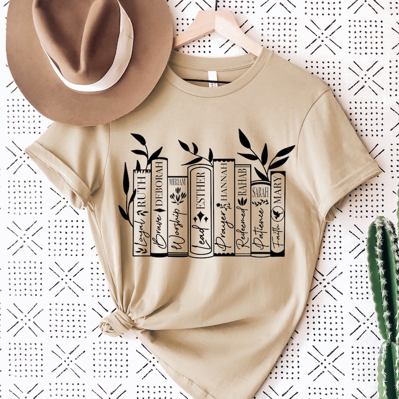 May include: A beige t-shirt with a black graphic of a bookshelf with books that have names of women from the Bible. The books are labeled: Loyal Ruth, Brave Deborah, Worship Miriam, Lead Esther, Prayer Hannah, Redeemed Rahab, Patience Sarah, Faith Mary.
