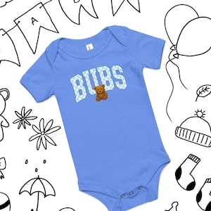 Puede incluir: Un body de bebé azul claro con la palabra "BUBS" en un patrón de cuadros y un pequeño oso de peluche. El body tiene mangas cortas y corchetes en la parte inferior. El fondo presenta dibujos en blanco y negro de globos y otras ilustraciones infantiles.