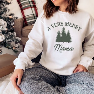 以下が含まれることがあります： 「A Very Merry Mama」という文字と、緑色の松の木3本のグラフィックがプリントされた白いスウェットシャツです。