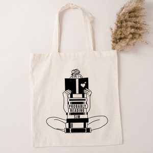 Könnte beinhalten: Eine weiße Einkaufstasche mit schwarzen Henkeln und einer schwarzen Illustration einer Person, die mit einem Stapel Bücher sitzt. Der Text auf der Tasche lautet "Probably Reading SJM".