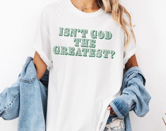 Puede incluir: Camiseta blanca con el texto verde "ISN'T GOD THE GREATEST?". La camiseta tiene mangas enrolladas y un corte relajado. Una chaqueta vaquera azul claro se lleva sobre los hombros.