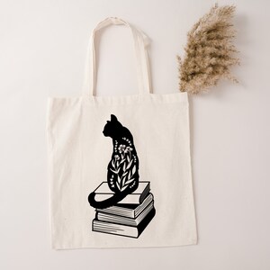 Puede incluir: Una bolsa de tela negra con la silueta de un gato sentado sobre una pila de libros. El gato está lleno de diseños florales.