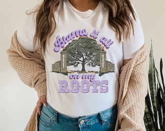 May include: White t-shirt with a purple and green graphic design that says "Blame it all on my roots". The graphic features a tree with two gates on either side.