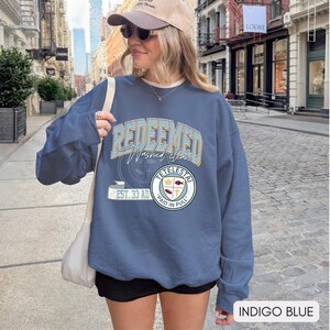 May include: Indigo blue sweatshirt with the words "REDEEMED Washed clean" in a collegiate style font. The sweatshirt also features a circular emblem with the words "TETELESTAI" and "PAID IN FULL". The text "EST. 33 AD" is also visible.