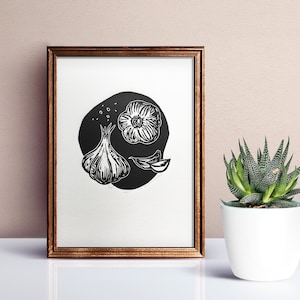 May include: A black and white linocut print of garlic cloves and a garlic bulb in a circle. The print is framed in a brown wood frame.