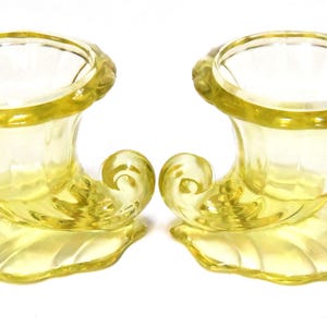 May include: A pair of yellow glass candle holders with a horn-like handle and a scalloped base.