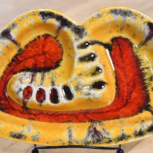 May include: A yellow ceramic ashtray with a red and black abstract design. The ashtray has three indentations for cigarettes.