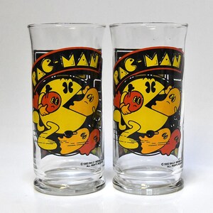 May include: Two clear glass drinking glasses with a Pac-Man design. The glasses feature a yellow Pac-Man eating a red ghost with three other ghosts in the background. The text "PAC-MAN" is printed in red and black on the front of each glass. The copyright information "©1982 BALLY MIDWAY MFG. CO. ALL RIGHTS RESERVED" is printed on the bottom of each glass.