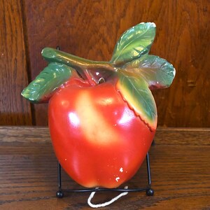 May include: A red ceramic apple with green leaves, on a black metal stand. The apple has a glossy finish and appears to be vintage.