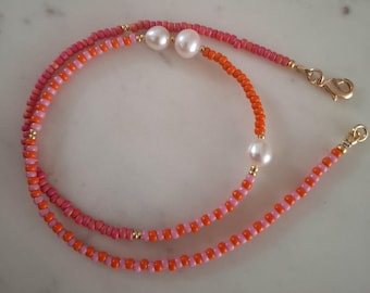 Puede incluir: Un collar de cuentas con una mezcla de cuentas rosas y naranjas. El collar presenta dos perlas blancas grandes y cierres dorados. Las cuentas son pequeñas y están espaciadas uniformemente, creando un diseño delicado.