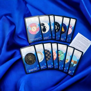 Op de afbeelding: Een set orakelkaarten met diverse illustraties, waaronder een klok, een zwart schaap en maanfasen. De kaarten zijn gerangschikt op een blauwe stoffen achtergrond. Elke kaart heeft een uniek ontwerp en tekst.