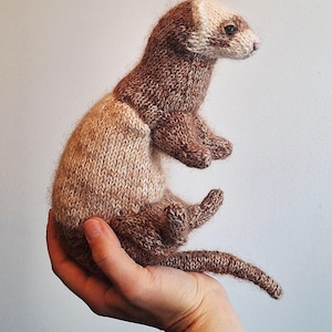 Op de afbeelding: Een handgebreid fret-speeltje met een tweekleurig ontwerp, met donkerbruine vacht op de kop, ledematen en staart, overgaand in een lichtere bruine tint op het middenstuk. De fret heeft een crèmekleurig gezicht en kleine donkere oogjes.