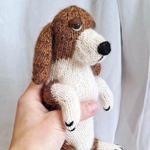 May include: A brown and white knitted dog toy with black eyes and nose. The toy is being held in a hand.