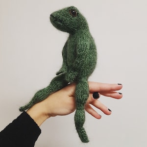 May include: A handmade, green knitted frog doll with large, brown eyes. The frog has long legs and arms, and is sitting upright. The doll is made of a textured yarn and has a soft, plush appearance. The background is plain white.