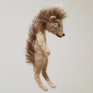May include: A brown knitted hedgehog toy with a white spiky back. The hedgehog is standing on its hind legs with its front legs tucked in.