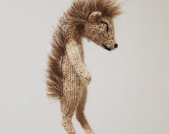 May include: A brown knitted hedgehog toy with a white spiky back. The hedgehog is standing on its hind legs with its front legs tucked in.