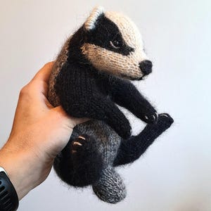Peut inclure: Un jouet blaireau tricoté présentant un visage rayé noir et blanc caractéristique, des membres noirs et un corps gris. L'animal en peluche a une texture duveteuse, des yeux sombres et un petit nez noir, réalisé avec des coutures détaillées.