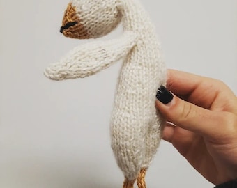 May include: A white knitted goose toy with a yellow beak and feet. The goose is standing on its feet with its wings outstretched.