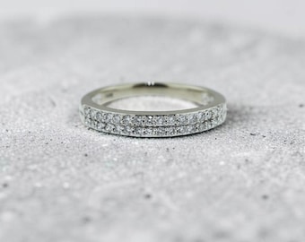 May include: A silver-toned metal band ring featuring a single row of small, clear, sparkling stones embedded along the outer surface. The ring has a polished finish and a slim, circular profile, showcasing a consistent design around the entire band.