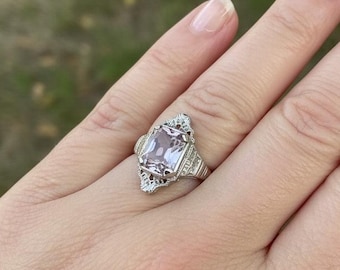 May include: A silver-toned ring featuring a rectangular, light pink gemstone set in a decorative metal mounting with intricate filigree details along the band. The setting includes small, clear accent stones surrounding the central pink stone.