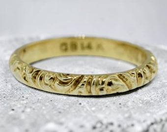 May include: A gold band ring featuring an intricate embossed scrollwork pattern along the exterior surface. The interior of the band is clearly stamped with the text GB14K, indicating the metal composition of the yellow gold ring.
