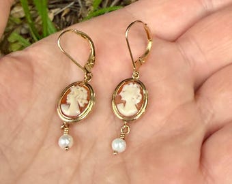 May include: A pair of gold-toned leverback earrings featuring oval-shaped cameos with a cream-colored profile relief on a reddish-brown background. Each earring is finished with a small, round, white pearl dangling from the bottom of the setting.
