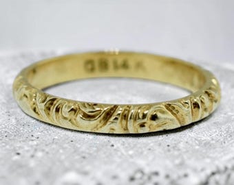 May include: A gold band ring featuring an intricate embossed scrollwork pattern along the outer surface. The interior of the ring is clearly stamped with the text GB 14K, indicating the metal composition and a maker mark on the inner band.